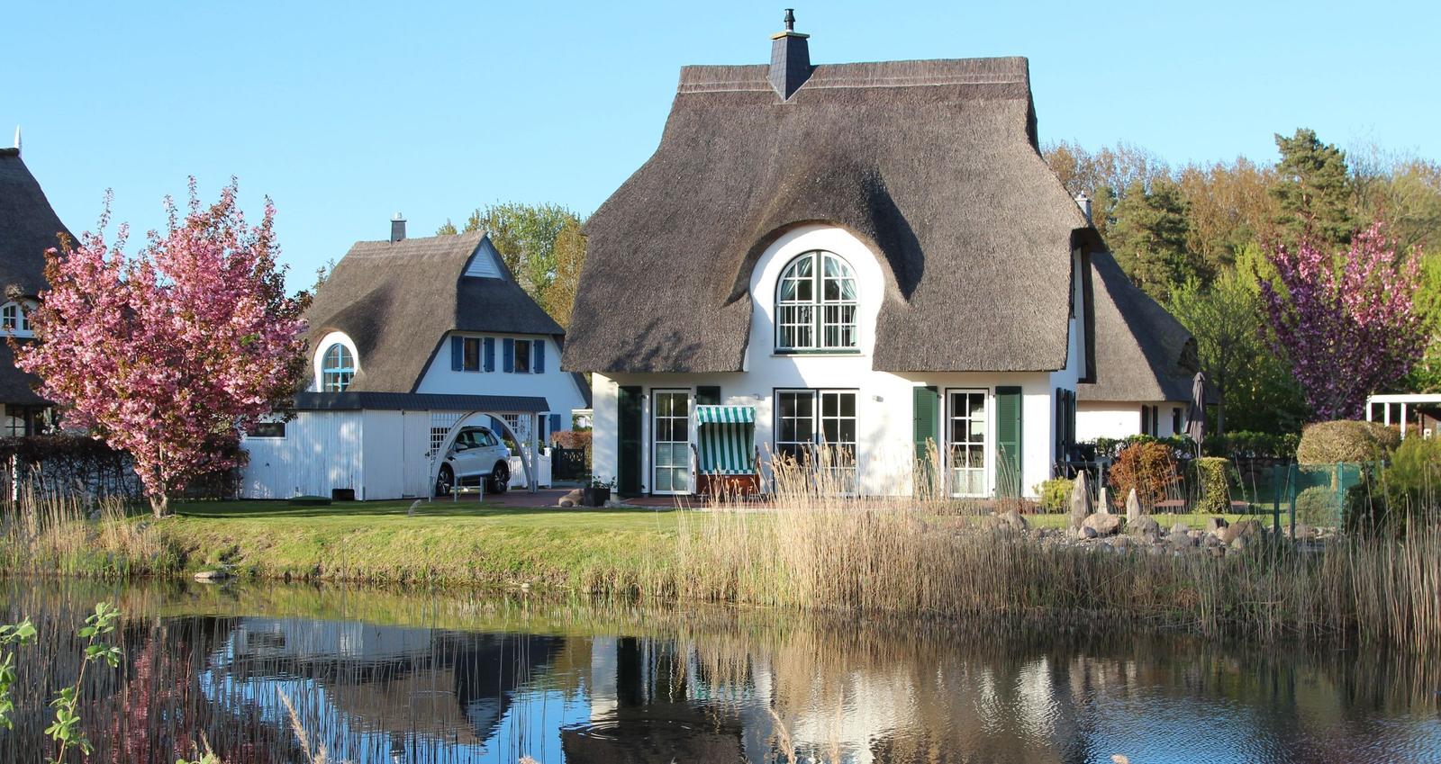 Haus mit Strohdach am Wasser, umgeben von Gras und blühenden Bäumen.