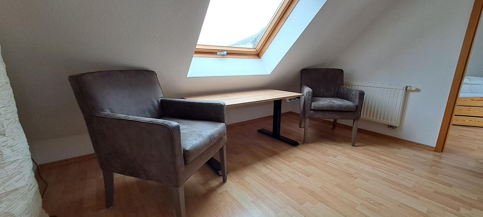 Zimmer mit Schreibtisch, zwei Stühlen und Dachfenster. Holzfußboden und Heizkörper sichtbar.