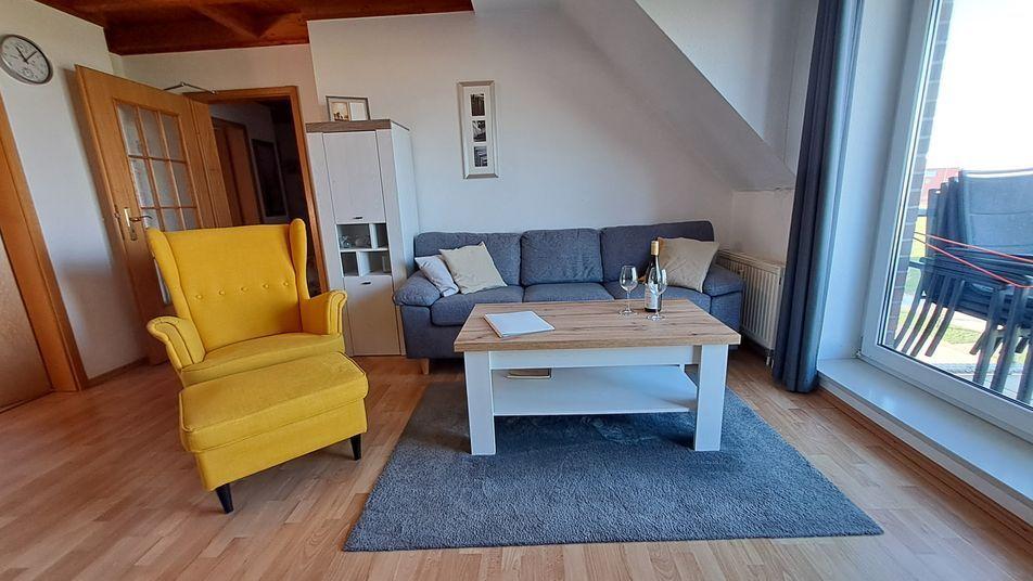 Wohnzimmer mit grauem Sofa, gelber Sessel und Holztisch. Große Fenster und Holzboden.
