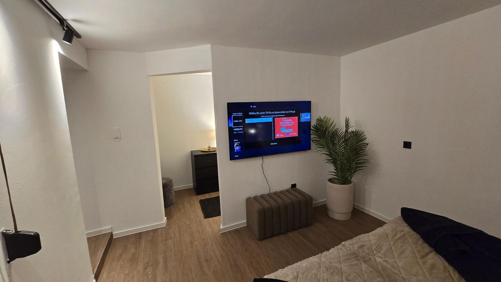 Schlafzimmer mit TV, Pflanze und Holzboden. Durchgang zur Nachbarwohnung.