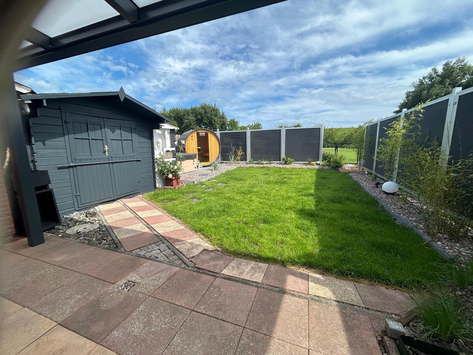 Hinterhof mit Rasen, Steinweg und Holzsauna. Umzäunt mit dunklem Holzzaun.