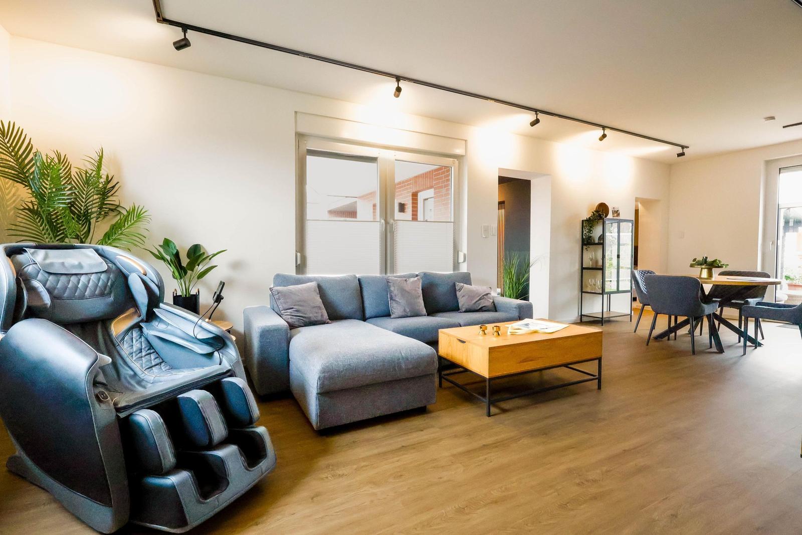 Wohnbereich mit Sofa, Massagestuhl und Essbereich. Holzboden und große Fenster.