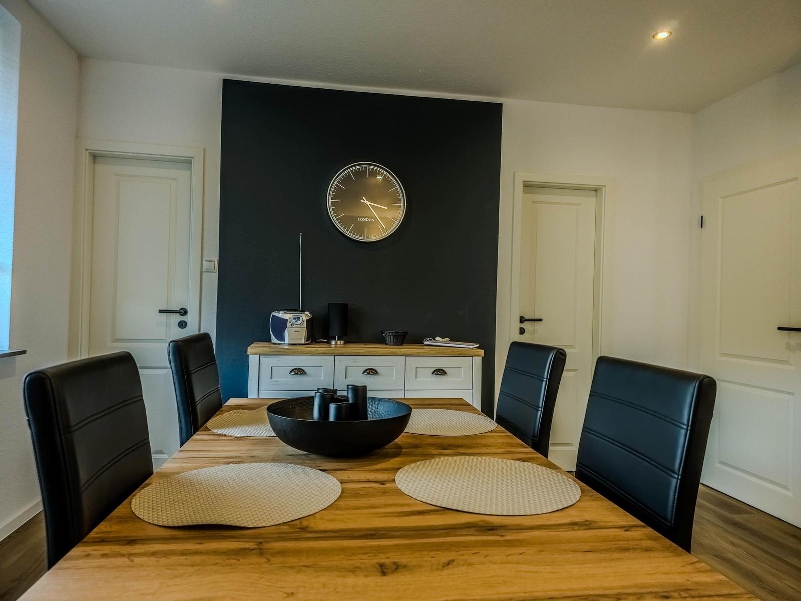 Dining room with wooden table, black chairs, and dark wall with clock.