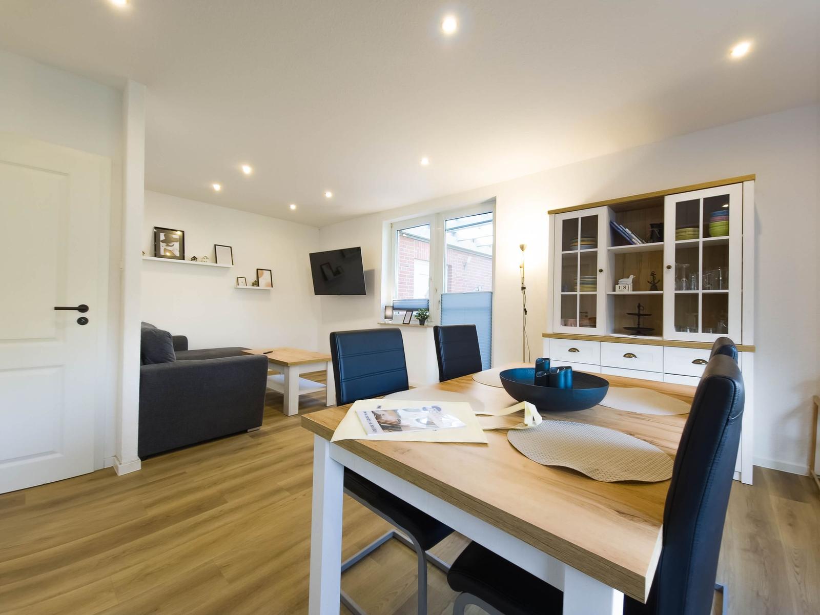 Dining area with wooden table and black chairs. Living area with sofa and TV in background.