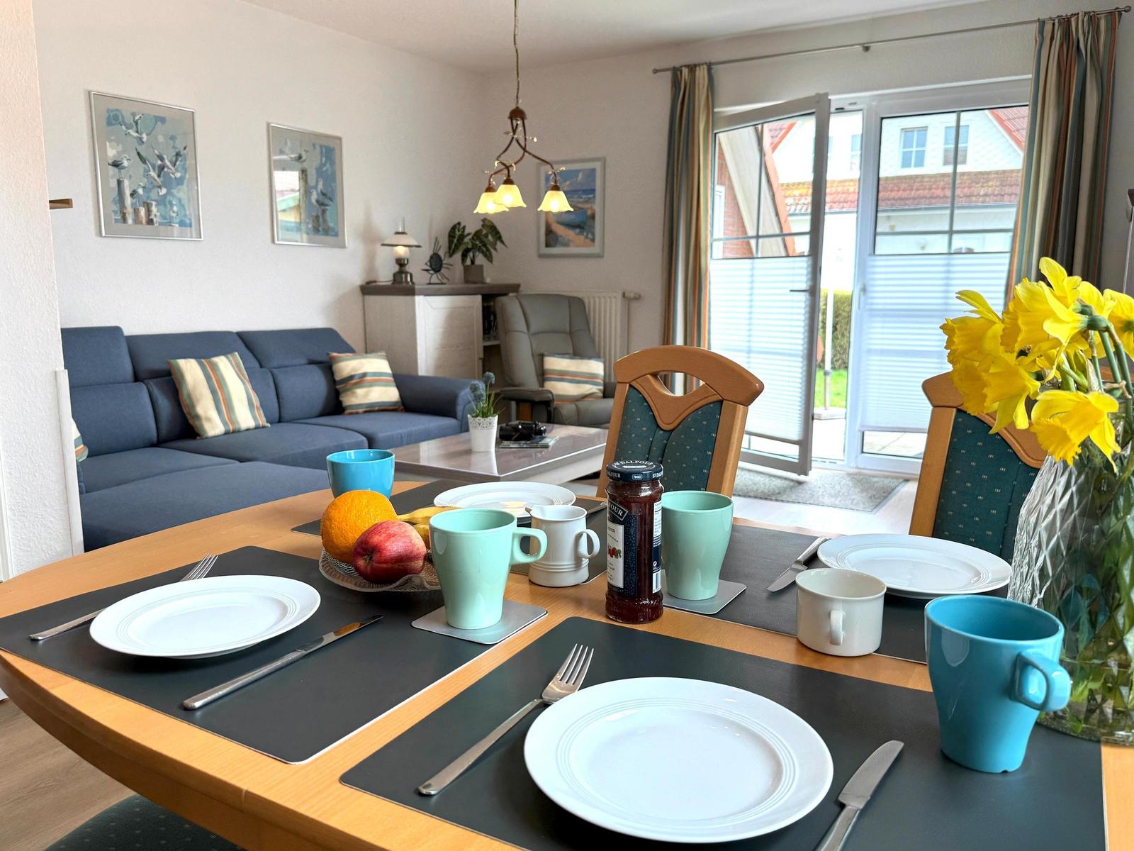 Esstisch mit Tischdecken, Tassen und Obst. Im Hintergrund Wohnzimmer mit Sofa und Glastür.