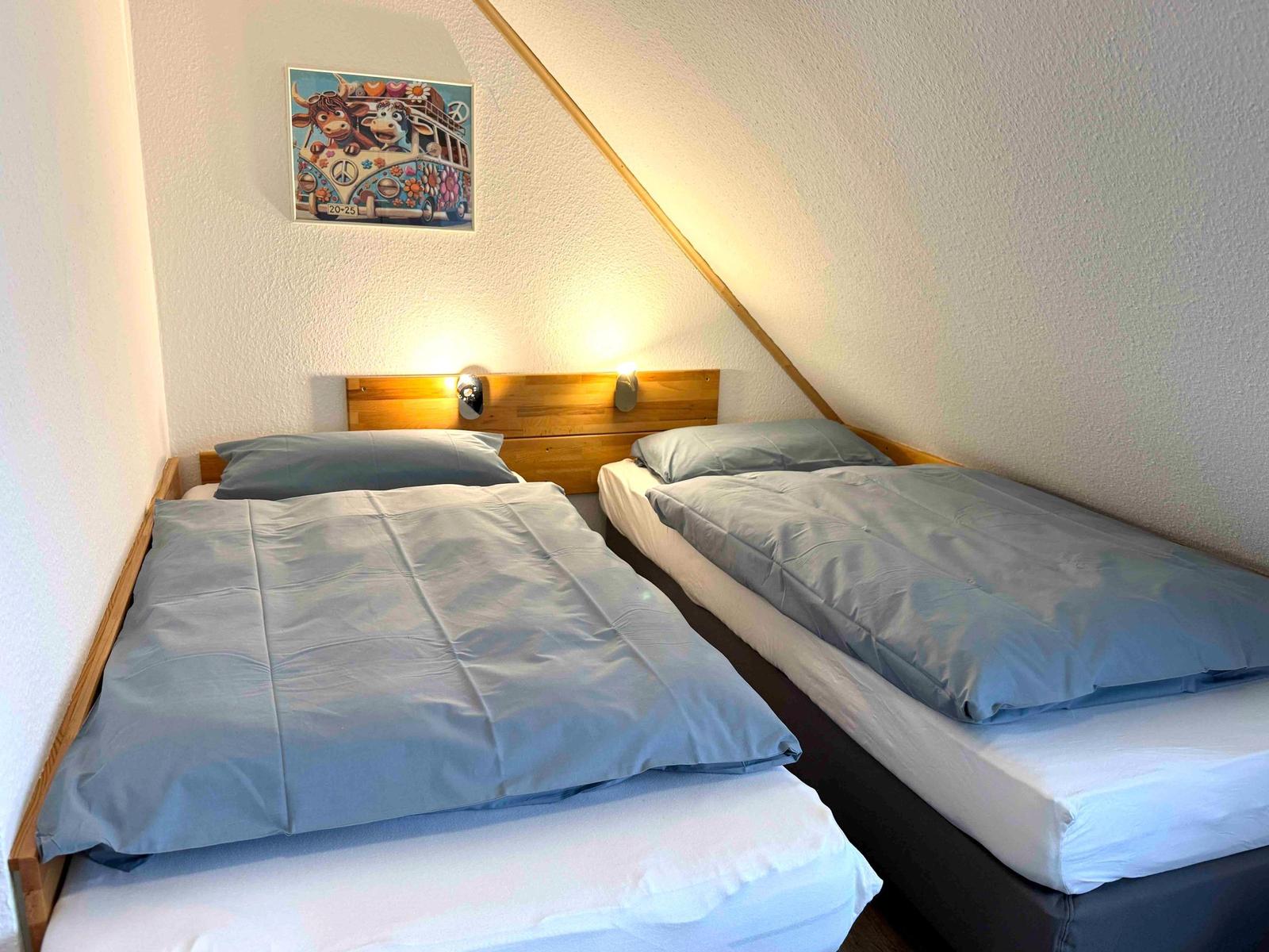 Zwei Einzelbetten mit Holzgestell und blauen Bettwäsche unter schräger Decke.