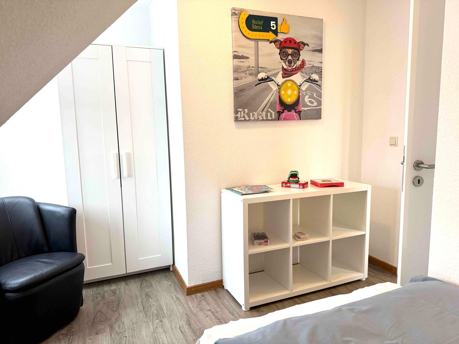 Schlafzimmer mit weißem Schrank, Bett und Regal. Wandbild zeigt einen Hund auf Motorrad.