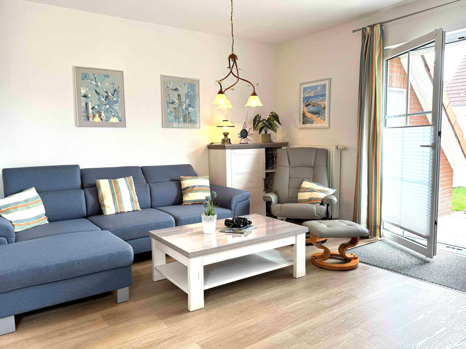 Wohnzimmer mit blauem Sofa, Couchtisch und Sessel. Fenster mit Blick nach draußen.