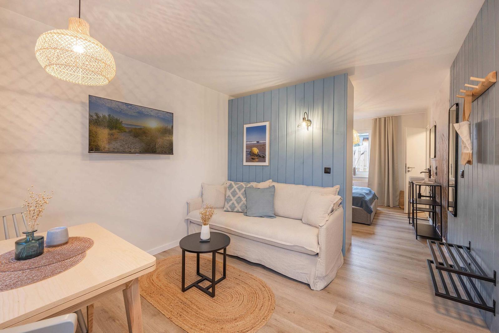 Wohnbereich mit Sofa, Esstisch und blauer Wandverkleidung. Blick in Schlafzimmer und Bad.