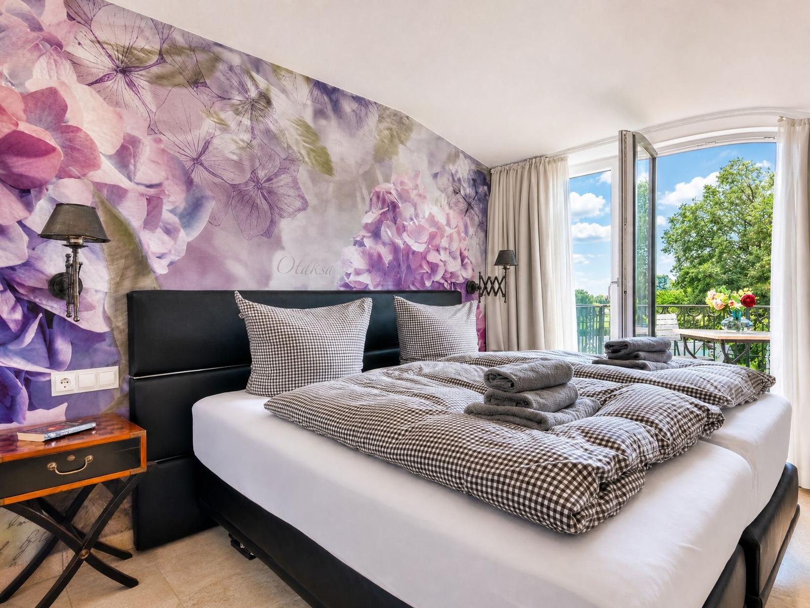 Schlafzimmer mit Blumenwand, Bett mit Kissen und Handtüchern, Balkon mit Sicht auf Grün.