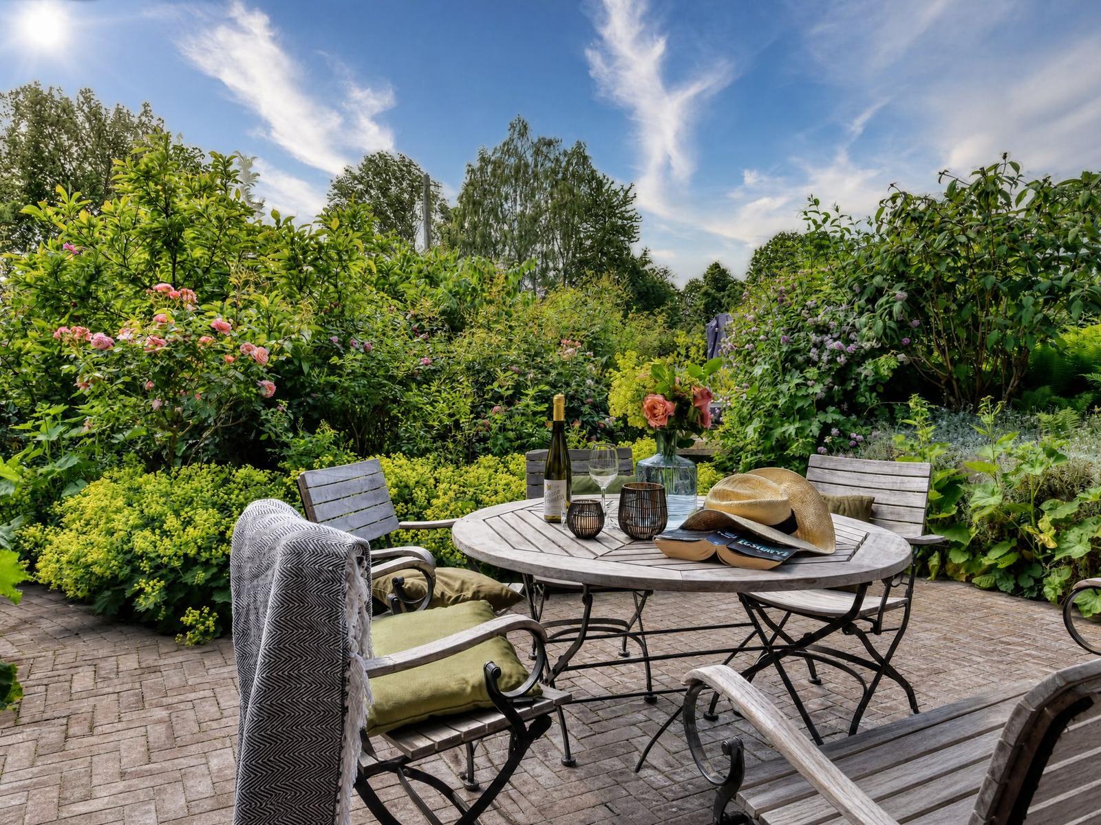 Terrasse mit Tisch, Stühlen und Blumenbeete im Garten. Flasche, Gläser und Hut darauf.