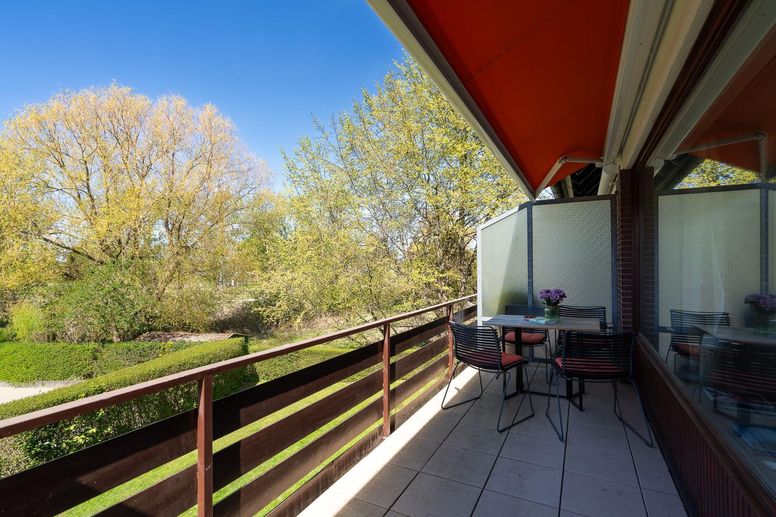 Terrasse mit Tisch, Stühlen und rotem Markisenüberdachung. Blick auf grüne Bäume und Hecken.