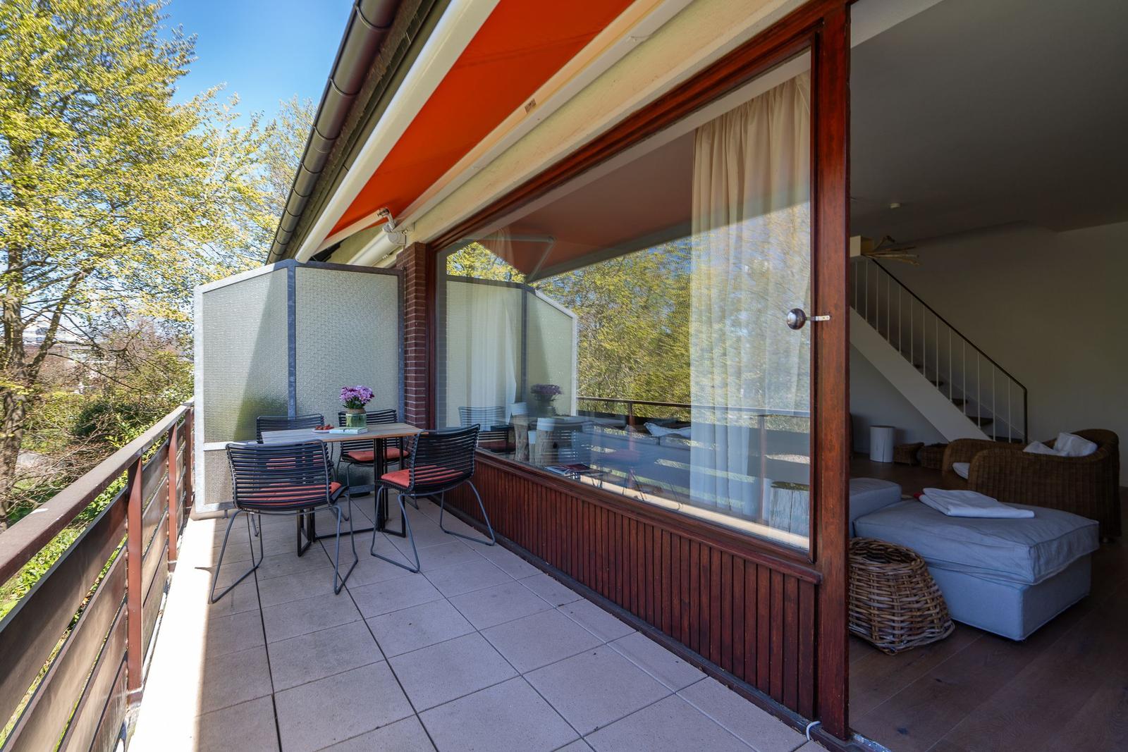 Terrasse mit Tisch, Stühlen und Sicht auf Bäume. Durchgehendes Glasfenster zum Innenraum.