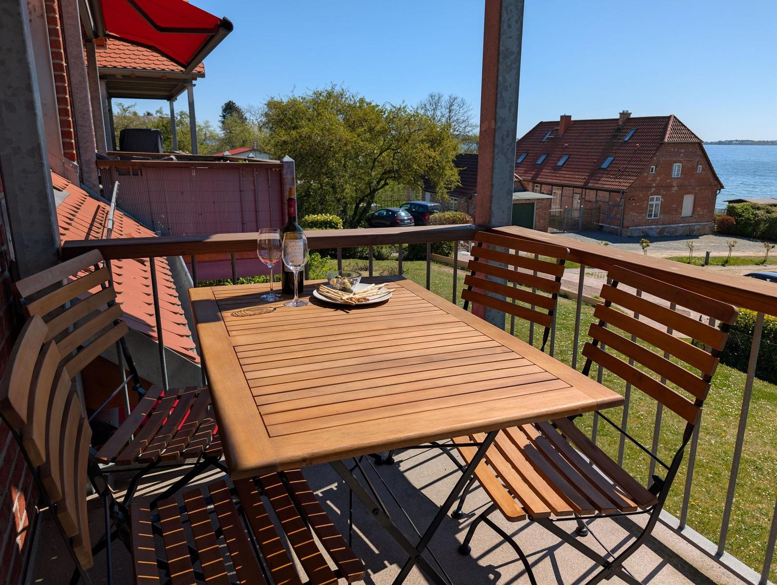 Terrasse mit Holztisch und Stühlen, Blick auf Haus und Wasser.