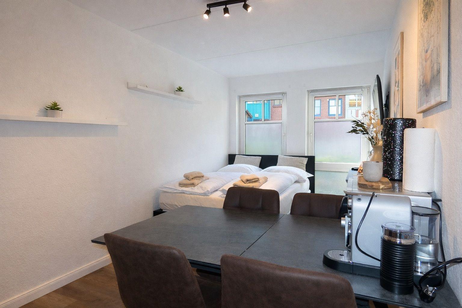 Schlafzimmer mit Bett, Tisch und Stühlen. Fenster und Wandregale sind sichtbar.