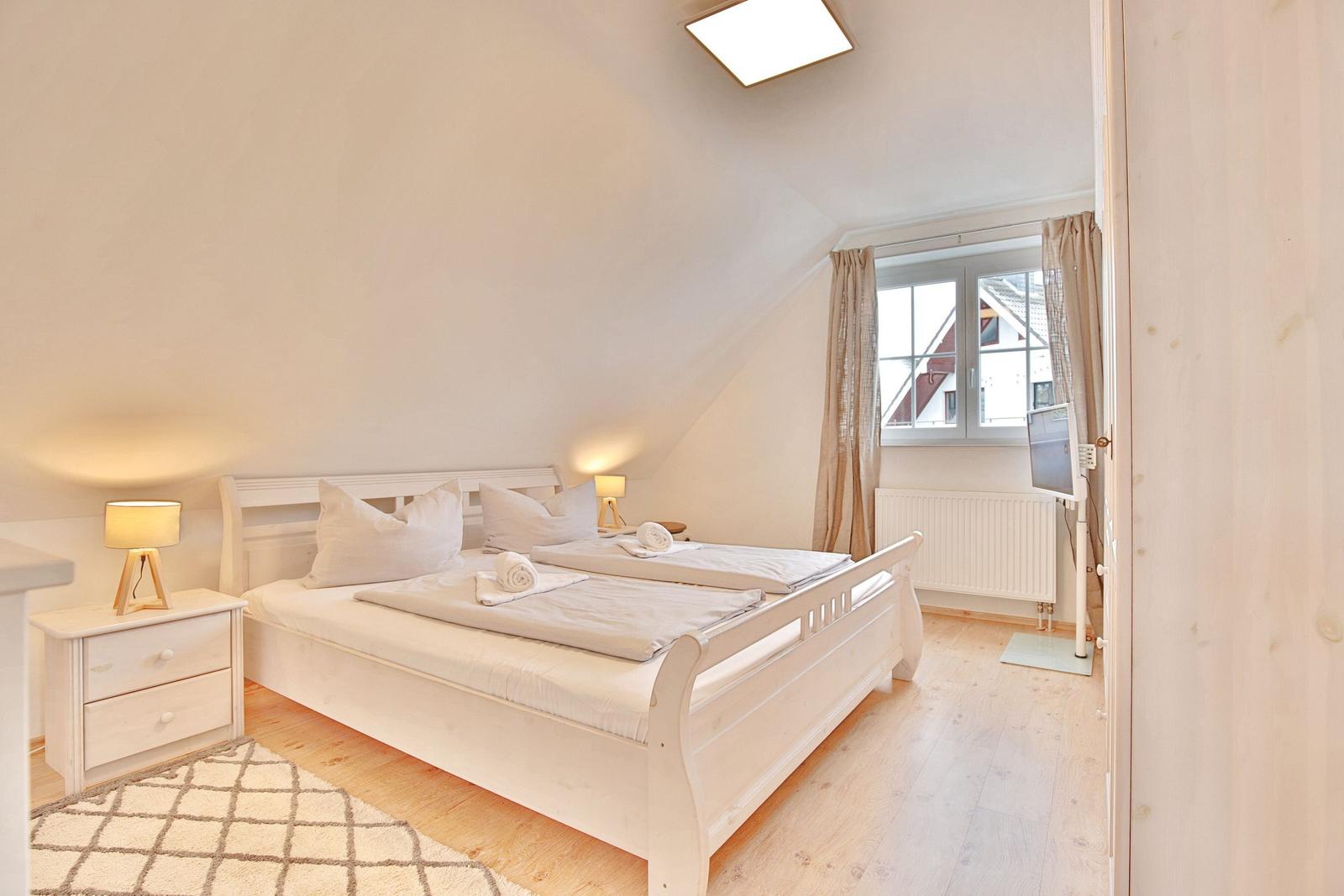 Schlafzimmer mit Doppelbett, Nachttisch und Fenster. Holzboden und Deckenleuchte.