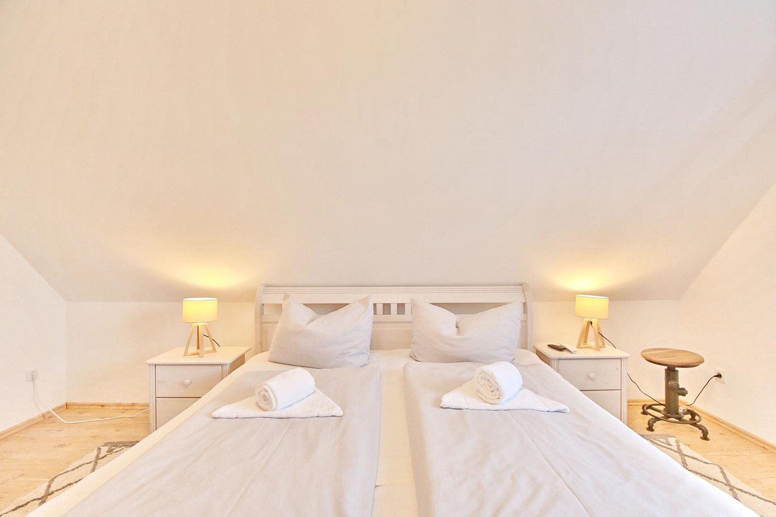 Zimmer mit Doppelbett, zwei Nachttischen und Lampen. Holzboden und schlichte Einrichtung.