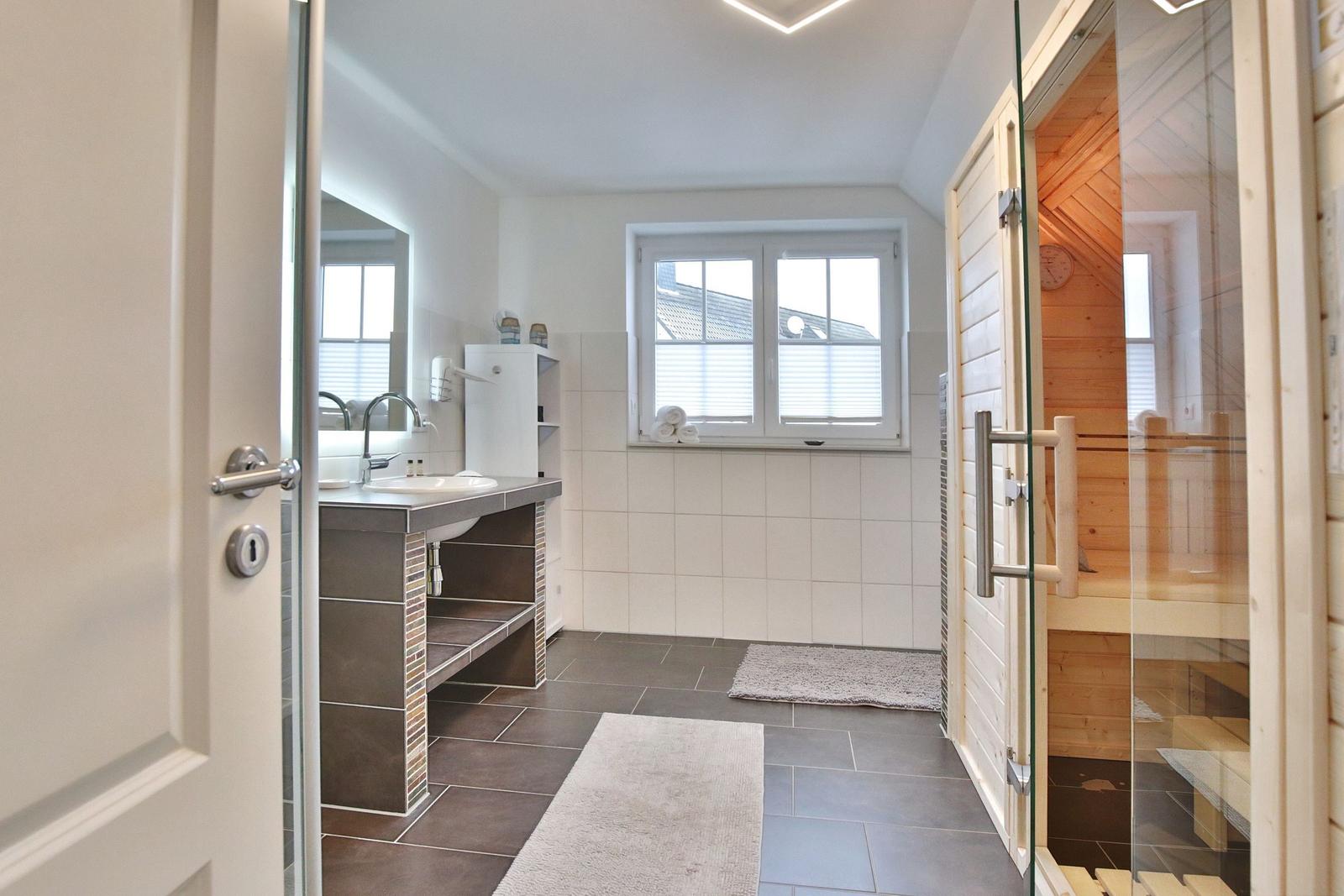 Badezimmer mit Dusche, Waschbecken und Sauna. Fenster mit Jalousie. Fliesenboden und Handtuch.