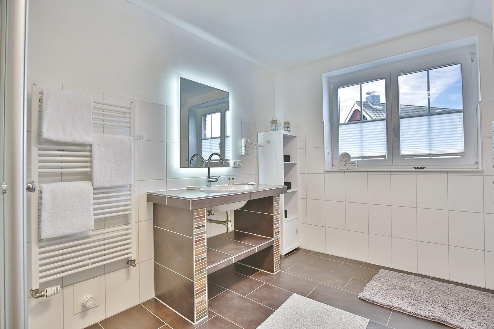 Badezimmer mit Waschbecken, Spiegel, Heizung und Fenster mit Jalousie.