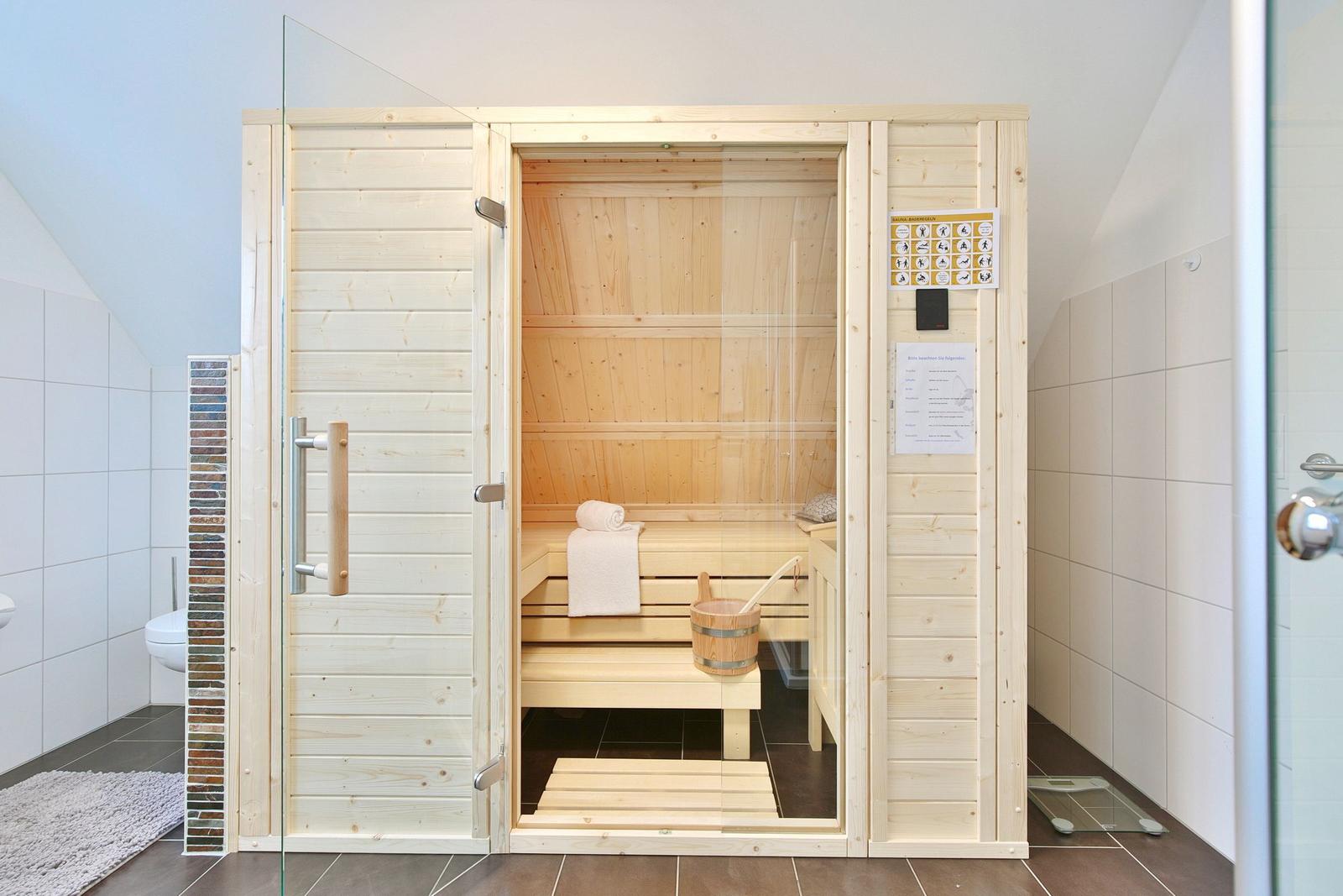 Holzsauna mit Sitzbank, Eimer und Handtuch. Glasfront und Anweisungstafel an der Wand.
