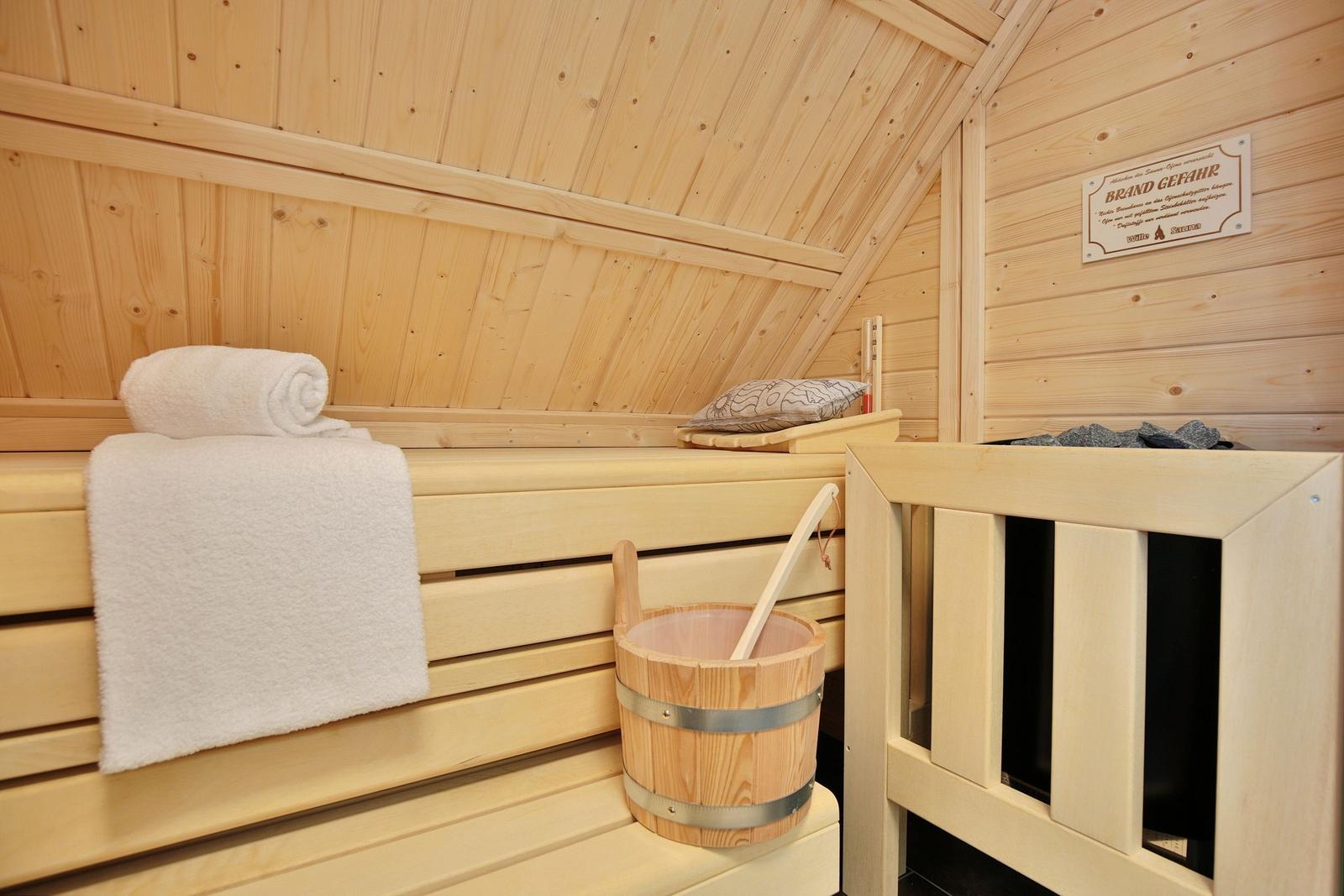 Sauna mit Holzbank, Handtuch und Eimer. Wandtafel mit Warnhinweis.