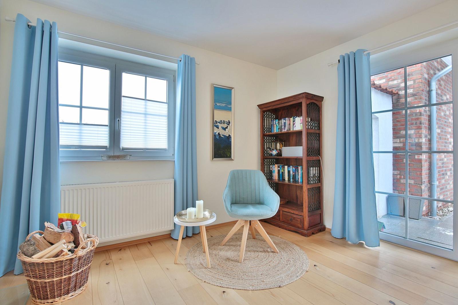 Ein Raum mit Holzboden, blauen Vorhängen, einem Bücherregal und einem Sessel.