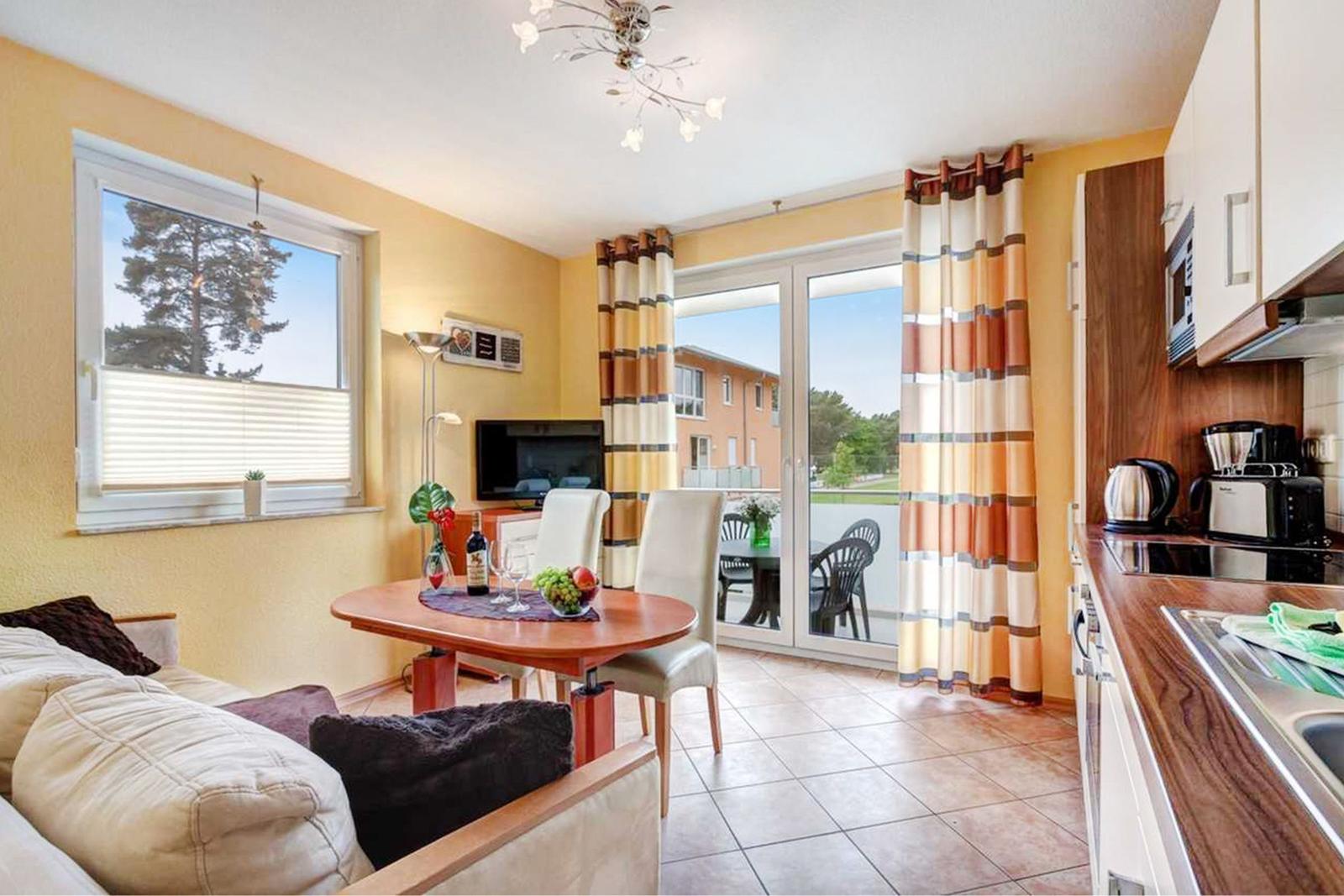 Wohn- und Essbereich mit Balkon, Küche und Fernseher. Holztisch, Sofa und Fenster mit Jalousie.