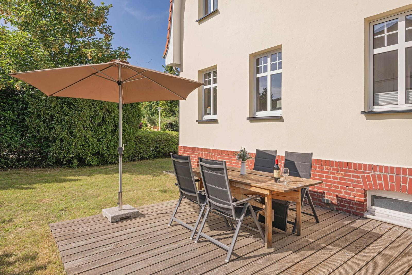 Deck with wooden table, four chairs, and umbrella. Background: house wall and greenery.