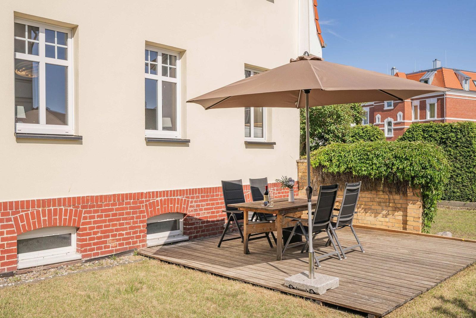 Deck with table, chairs, and umbrella. Background: house wall and green hedge.