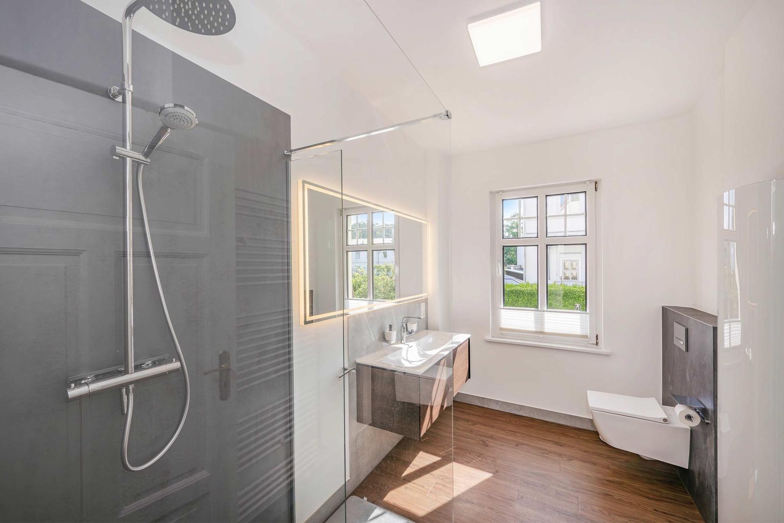 Modern bathroom with shower, sink, and toilet. Wooden floor and window with view.