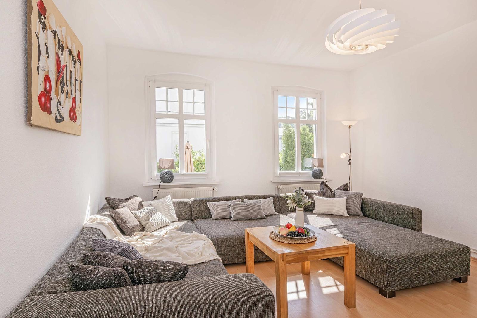 Living room with L-shaped sofa, wooden table, and two windows overlooking the garden.