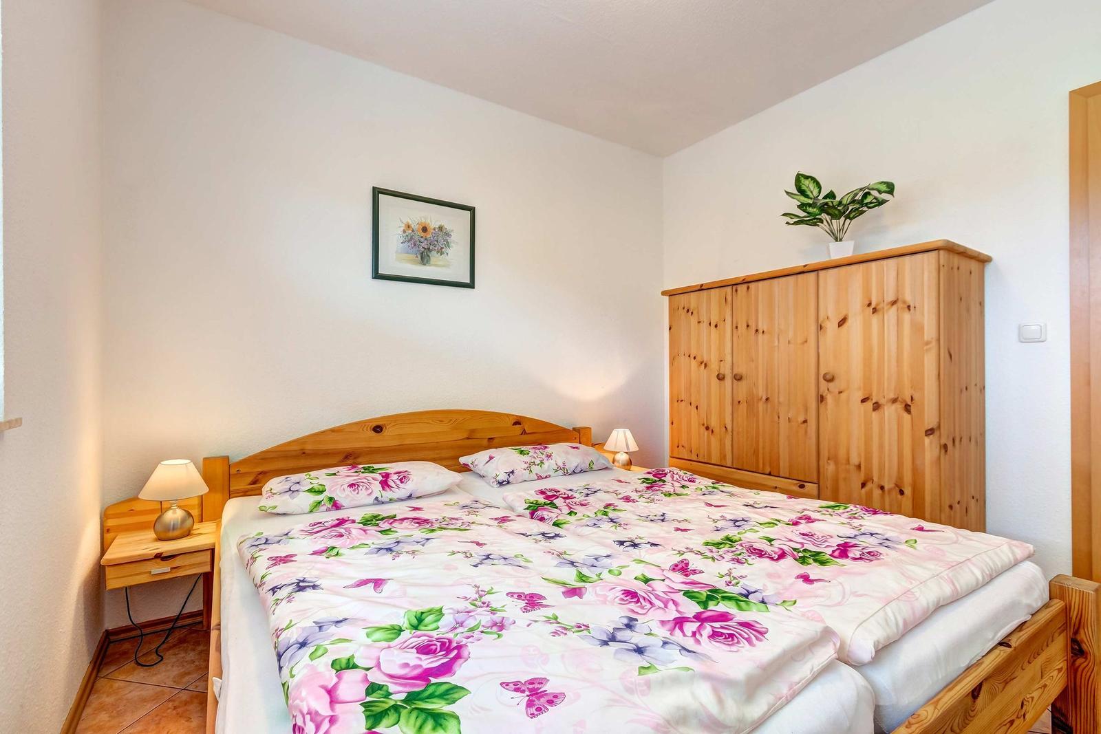 Schlafzimmer mit Holzbett, Nachttisch und Kleiderschrank. Bett mit floraler Bettwäsche.
