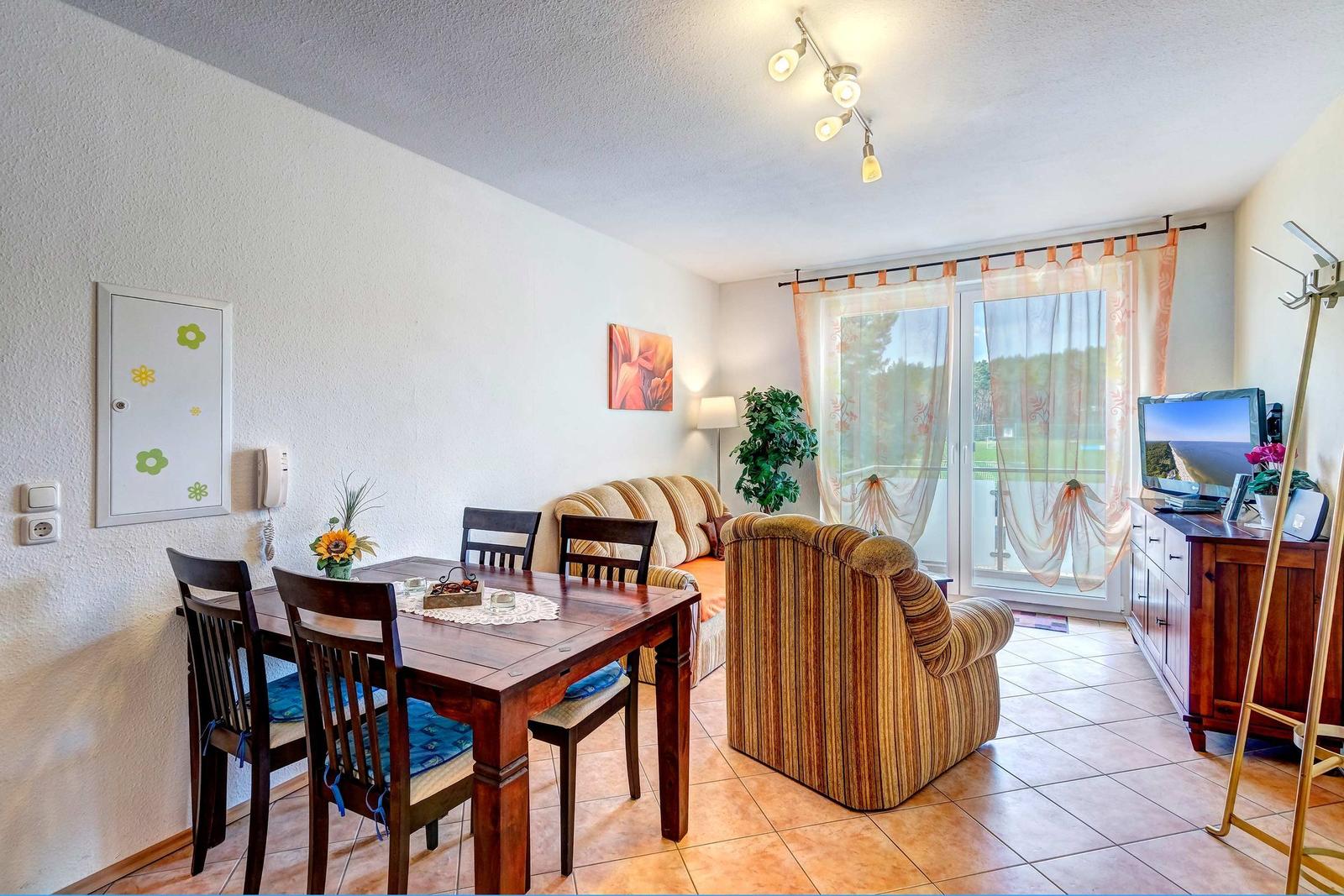 Wohn- und Essbereich mit Holztisch, Stühlen, Sofa und Fernseher. Terrasse mit Blick auf Grünfläche.