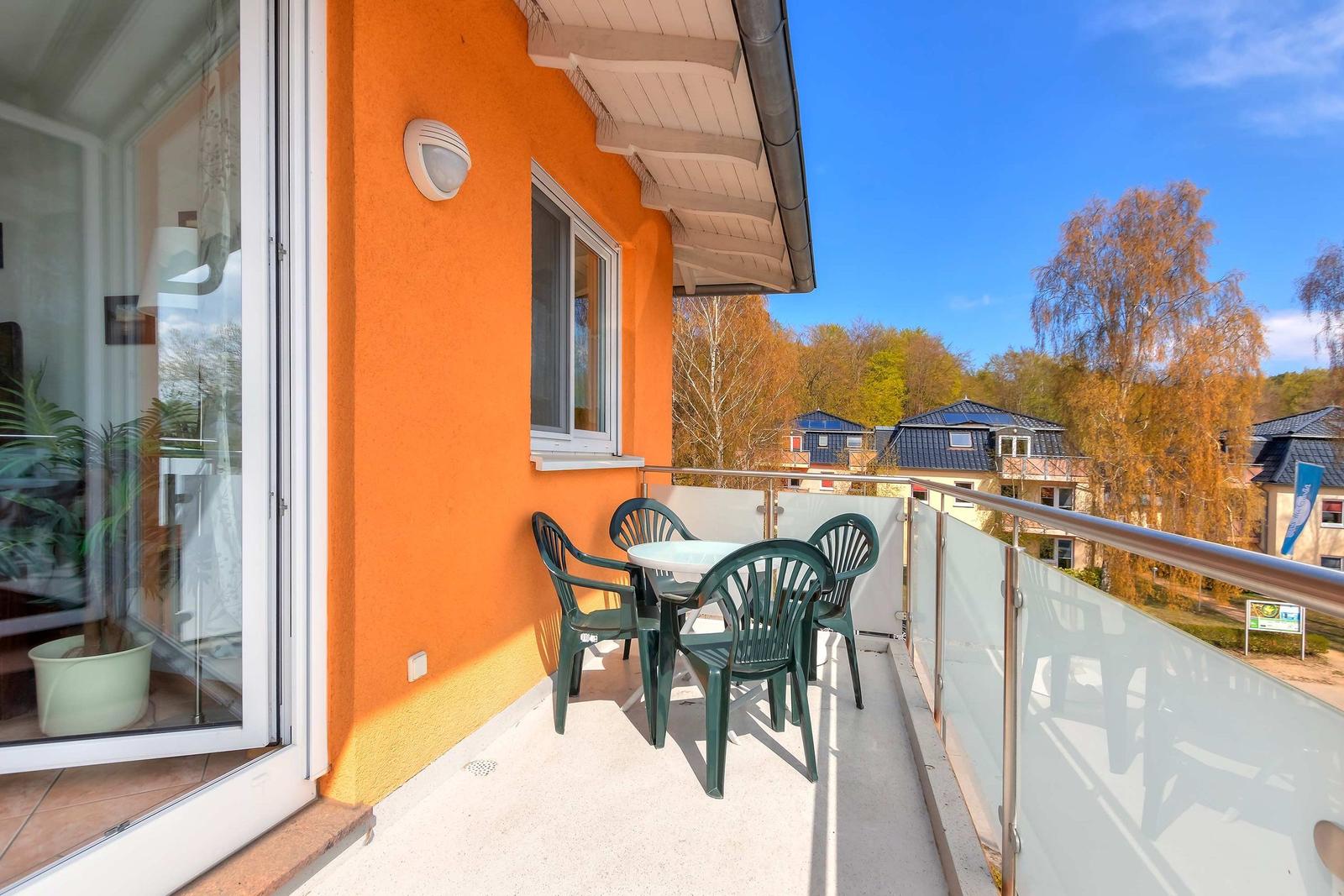 Balcony with green chairs and table, orange wall, glass railing, view of houses and trees.