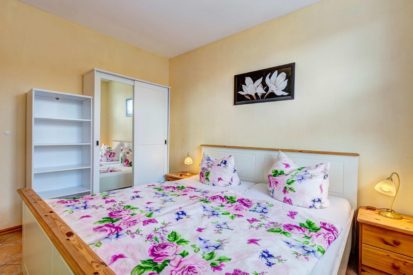 Schlafzimmer mit Bett, Nachttisch und Kleiderschrank. Wandbild und Blumenbezug.