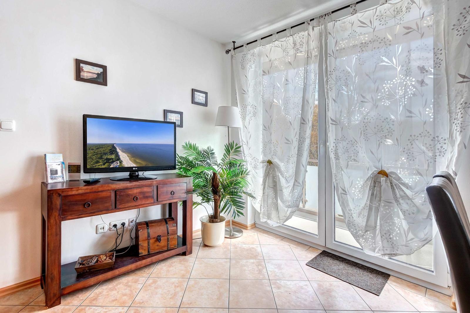 Wohnzimmer mit Fernseher, Pflanze und Balkontür mit Vorhängen.