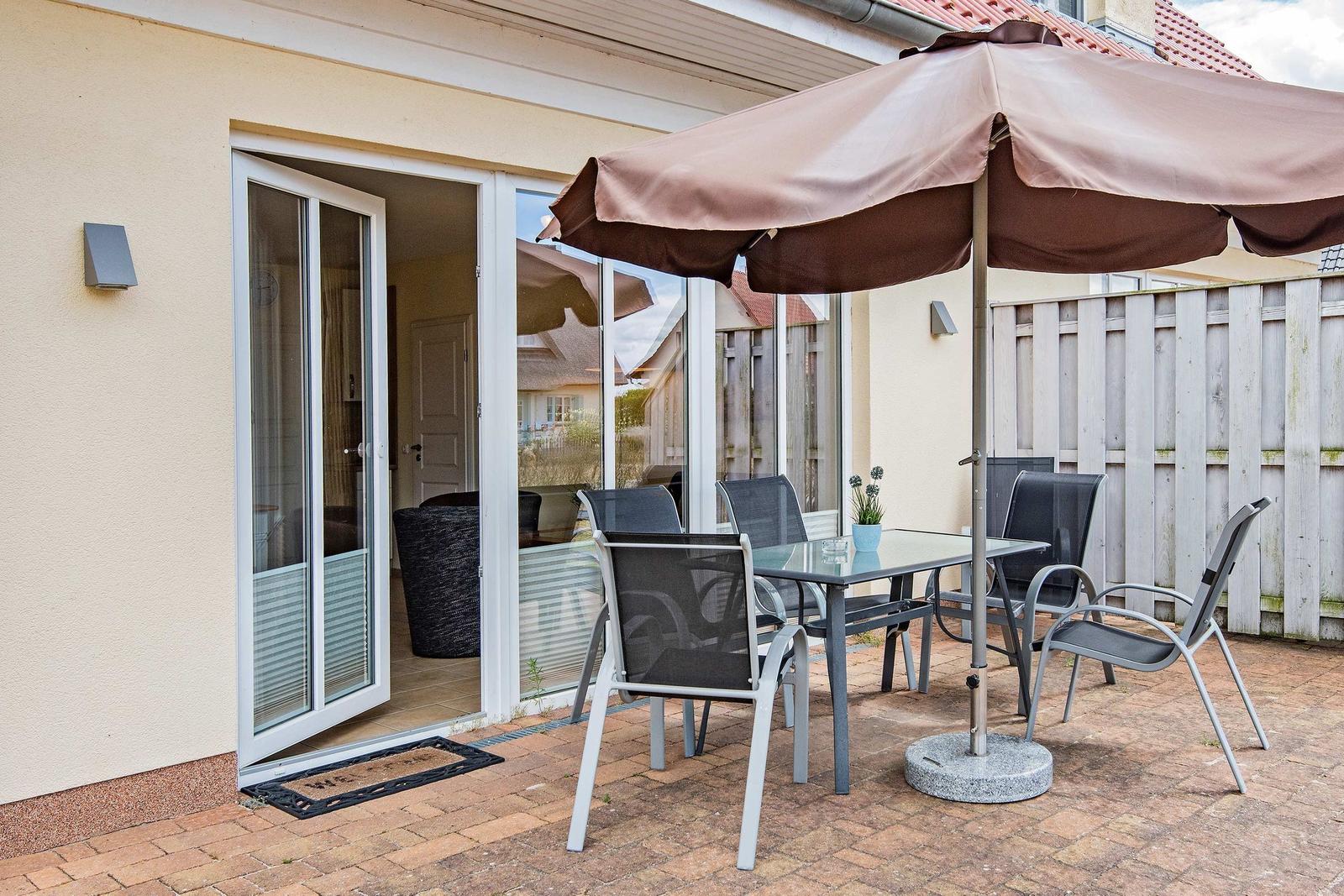 Terrasse mit Tisch, Stühlen und Sonnenschirm. Durchgang in das Haus. Holzzaun im Hintergrund.