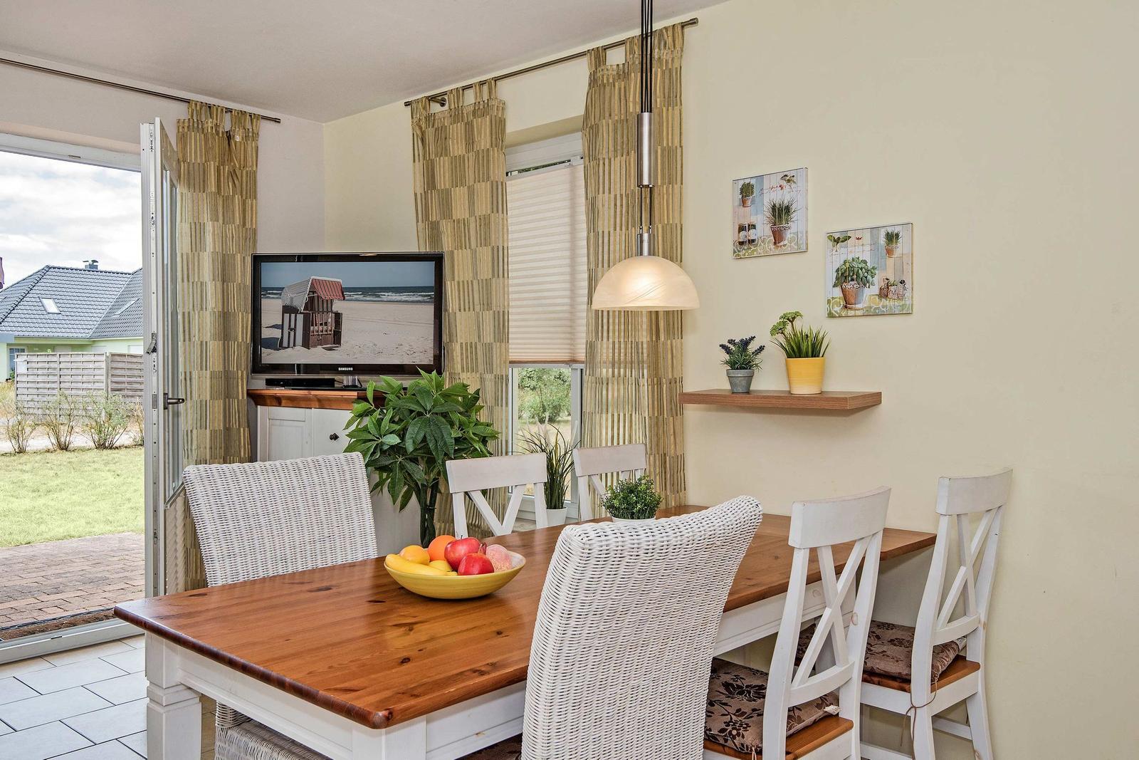 Esszimmer mit Holztisch, weißen Stühlen und Fernseher. Draußen liegt ein Garten.