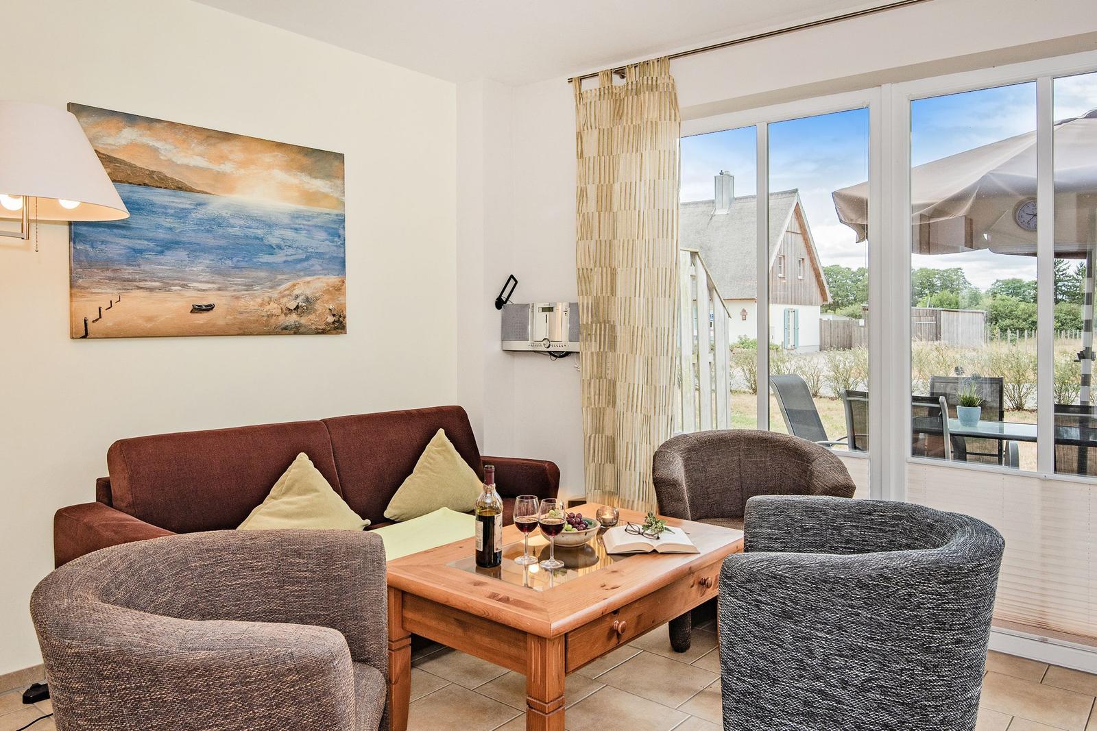 Wohnzimmer mit Sofa, Sesseln und Tisch. Durchgang zur Terrasse mit Gartenansicht.