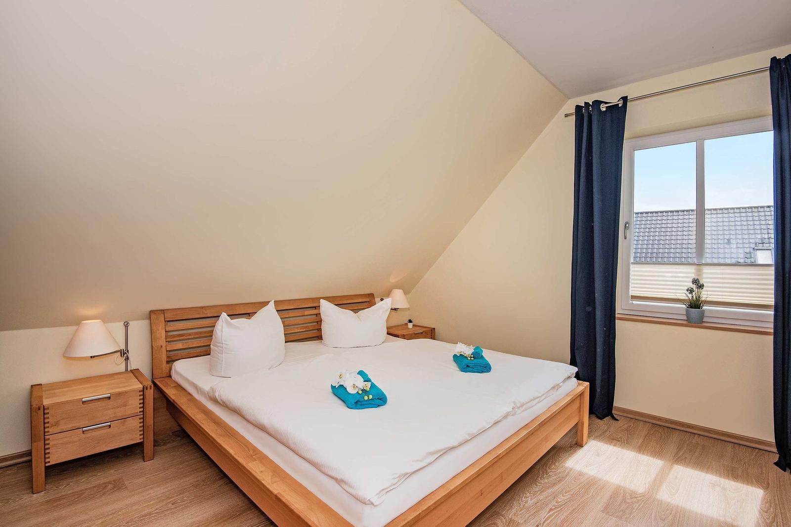 Doppelbett mit weißen Kissen und blauen Handtüchern. Holzfußboden und Fenster mit Vorhängen.