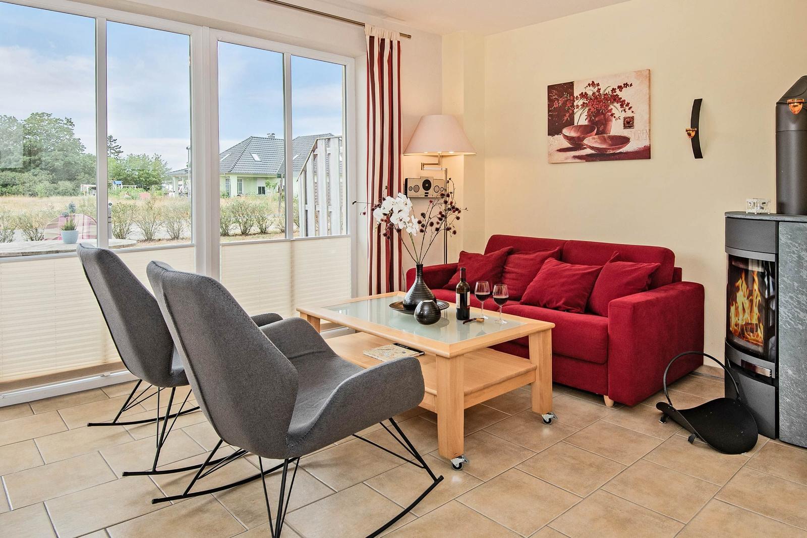 Ein Wohnzimmer mit rotem Sofa, grauen Stühlen und Kamin. Große Fenster bieten Blick auf Außenbereich.