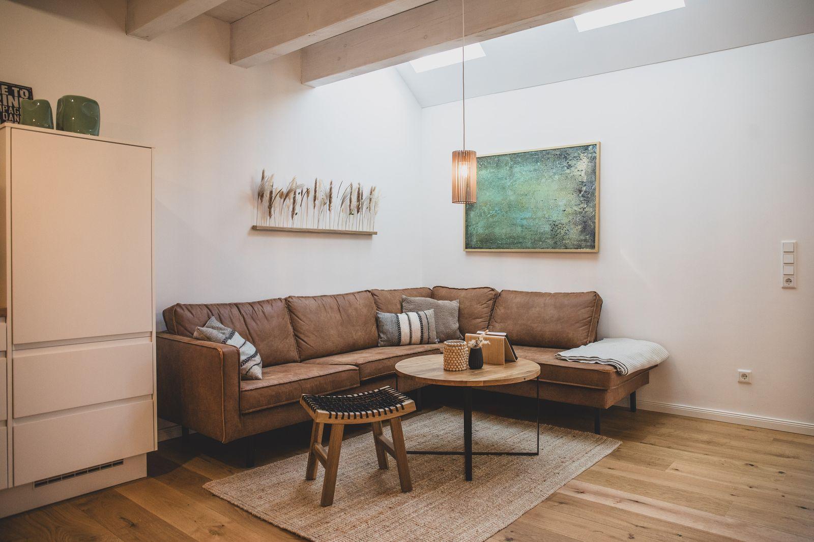 Ein Wohnzimmer mit braunem Sofa, Holzboden und Deckenbalken. An der Wand hängt ein Bild.