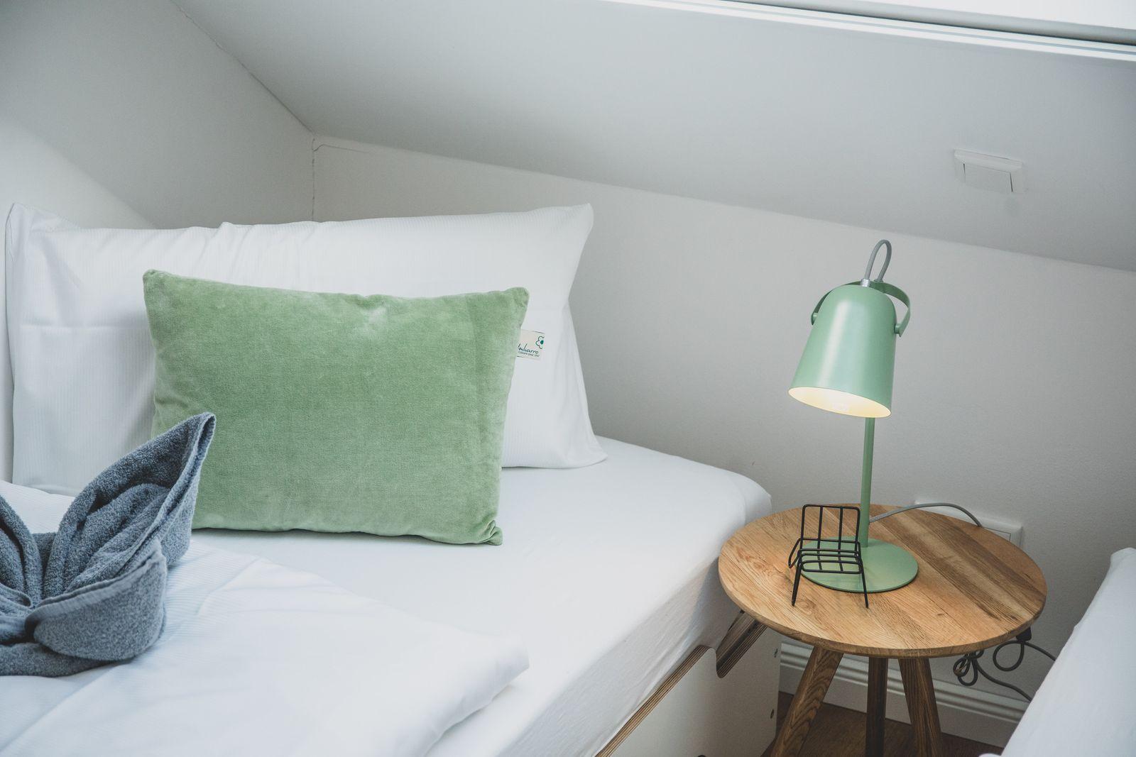 Ein Bett mit weißer Bettwäsche, grünem Kissen und einer Lampe auf einem runden Tisch.