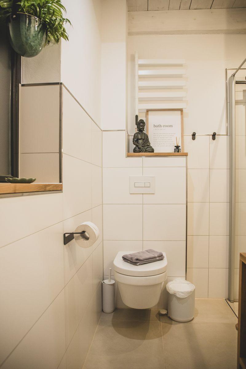 Badezimmer mit Toilette, Duschbereich und Pflanze. Buddha-Statue und Heizkörper sichtbar.