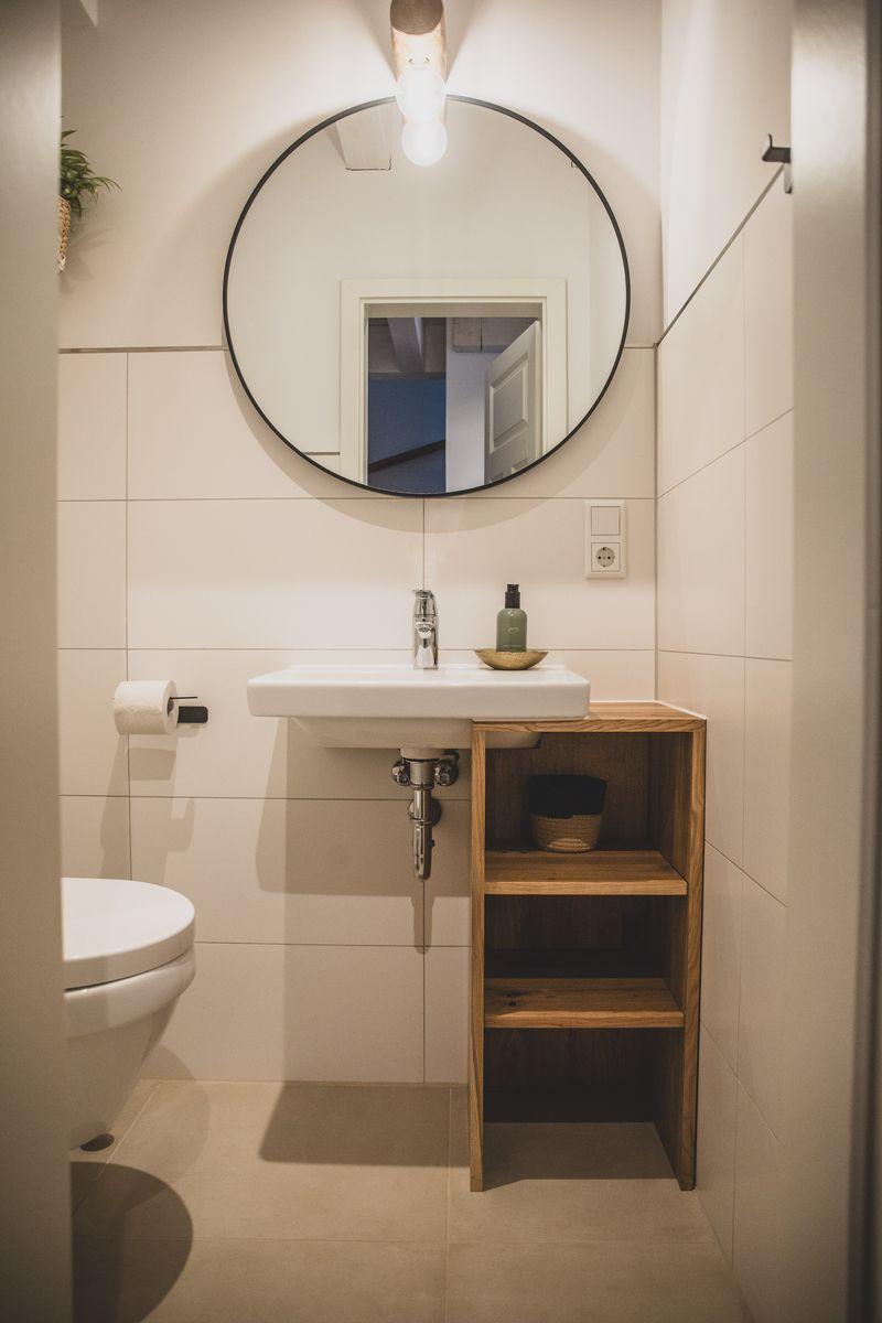 Badezimmer mit Waschbecken, Spiegel und Holzregal. Toilette und Duschwand sind sichtbar.