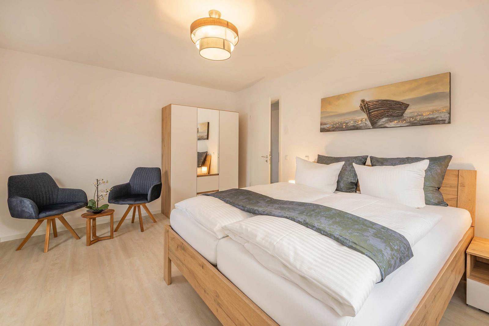 Schlafzimmer mit Bett, zwei Stühlen und Wandbild.