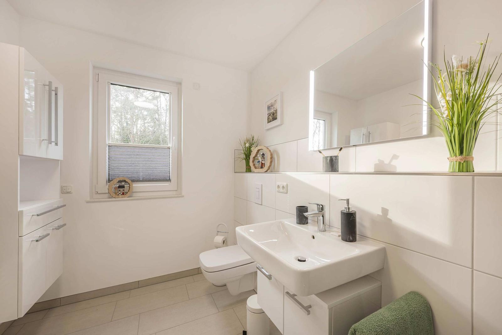 Badezimmer mit Waschbecken, Toilette und Fenster. Spiegelschrank und Pflanze auf der Arbeitsplatte.