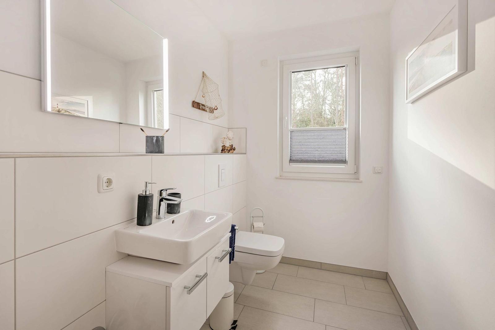 Badezimmer mit Waschbecken, Spiegel und Toilette. Fenster mit Jalousie.