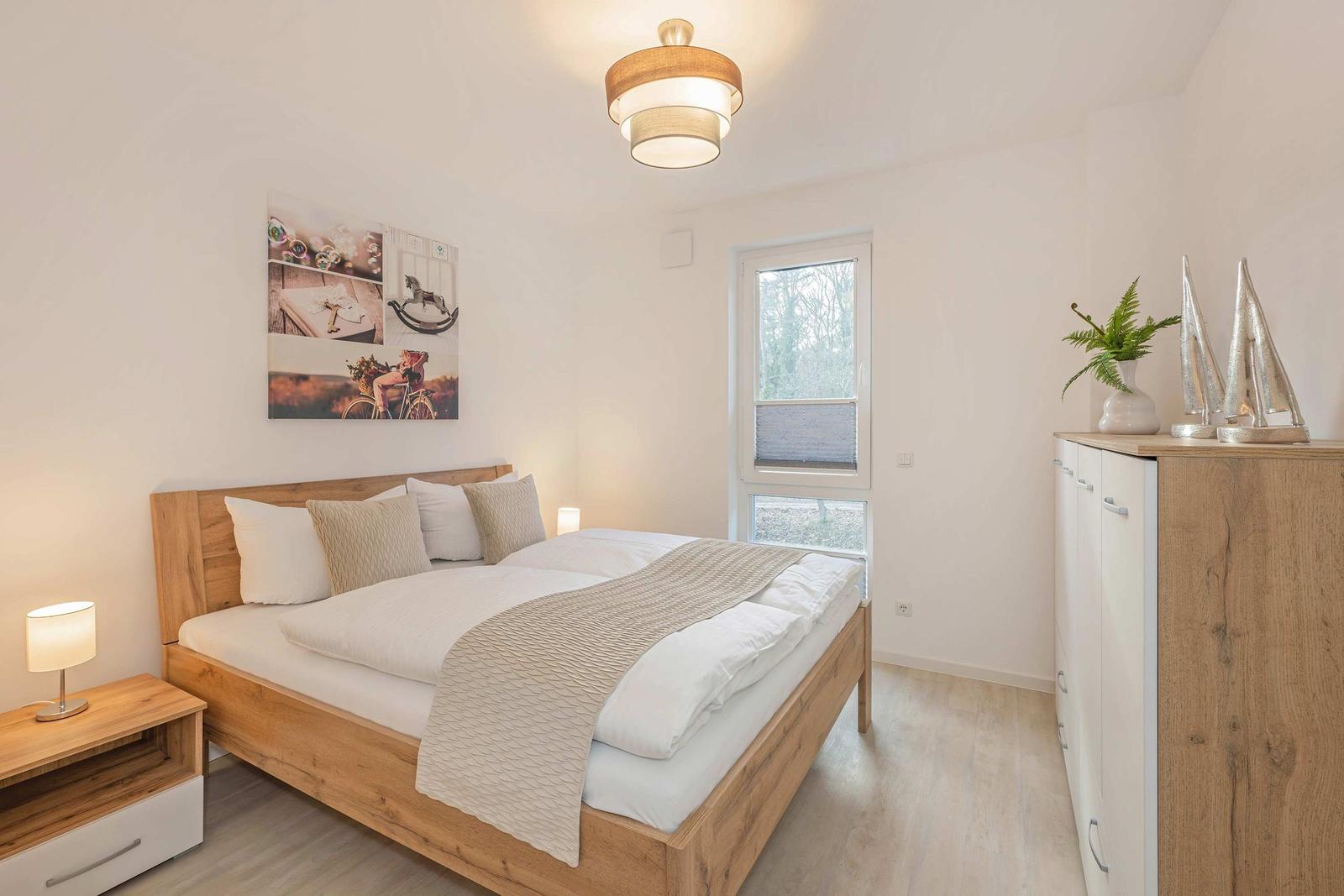 Schlafzimmer mit Holzbett, Nachttisch, Kleiderschrank und Fenster. Wandbild und Lampe sichtbar.
