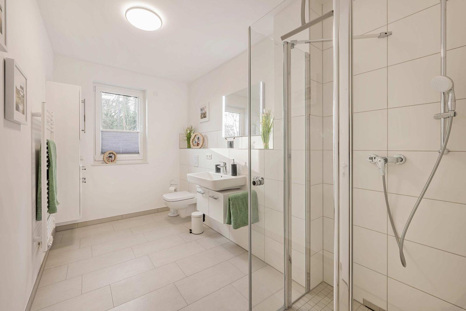 Badezimmer mit Dusche, Waschbecken und Toilette. Fenster, Spiegel und Handtuchhalter sichtbar.
